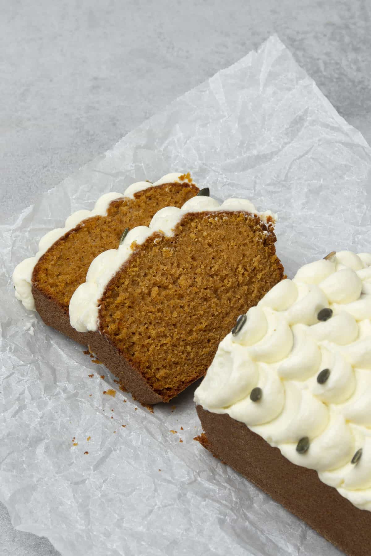 Pumpkin bread with cream cheese frosting on a white paper and two slices are lying on their side