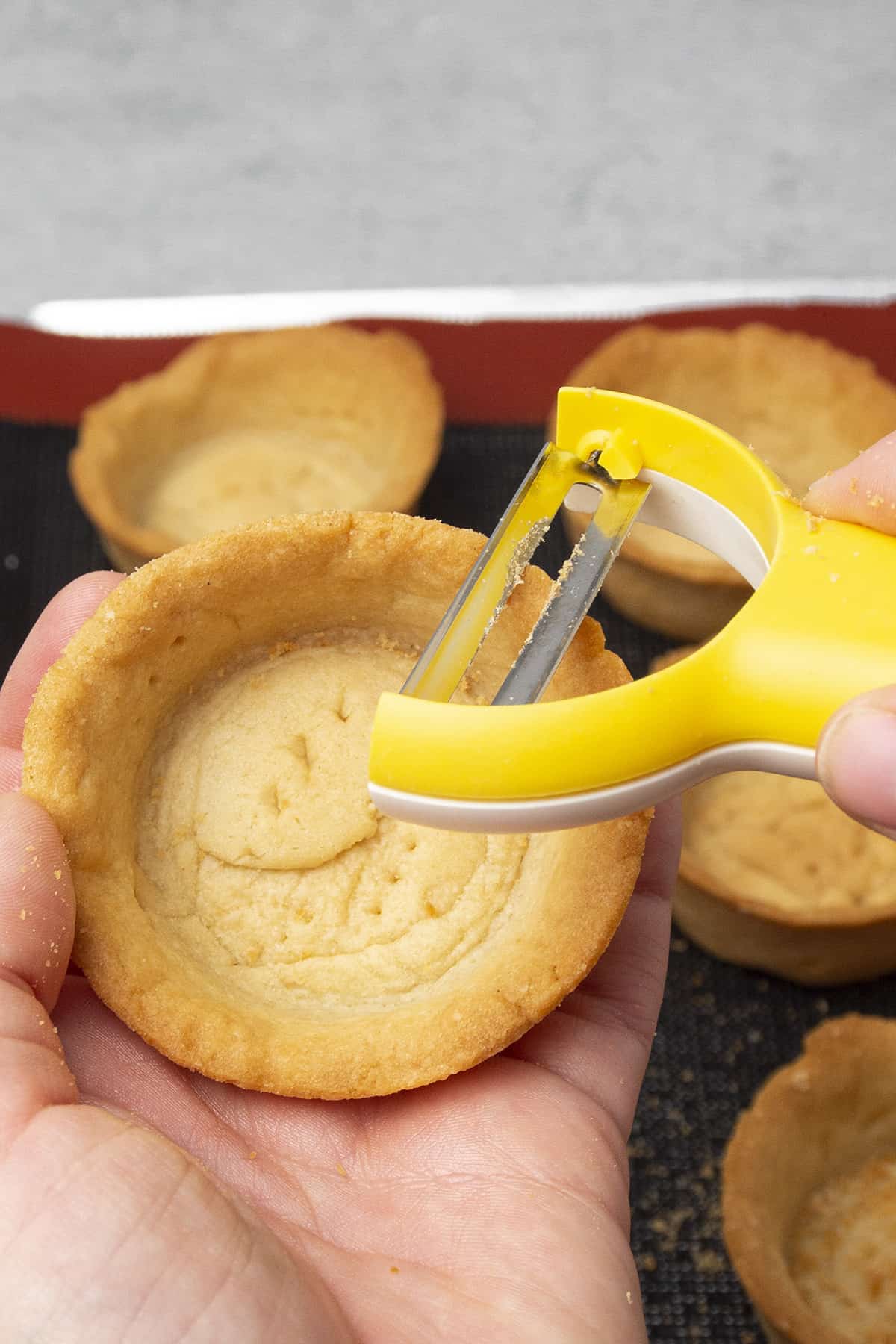 A hand holding a tart shell and sharpening the edge of the tart with a potato peeler