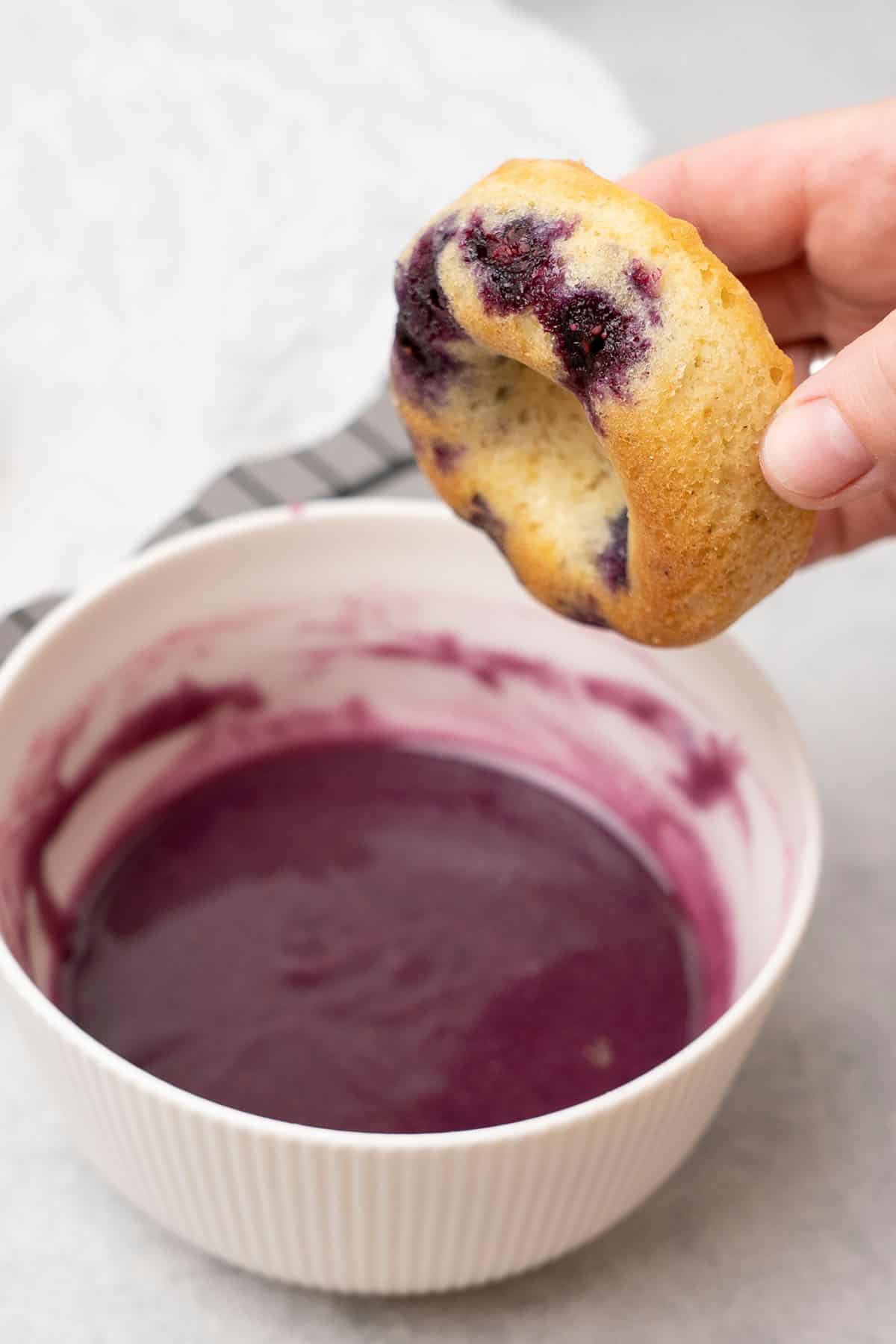 white ramekin filled with blueberry glazing and a hand dipping a donut.