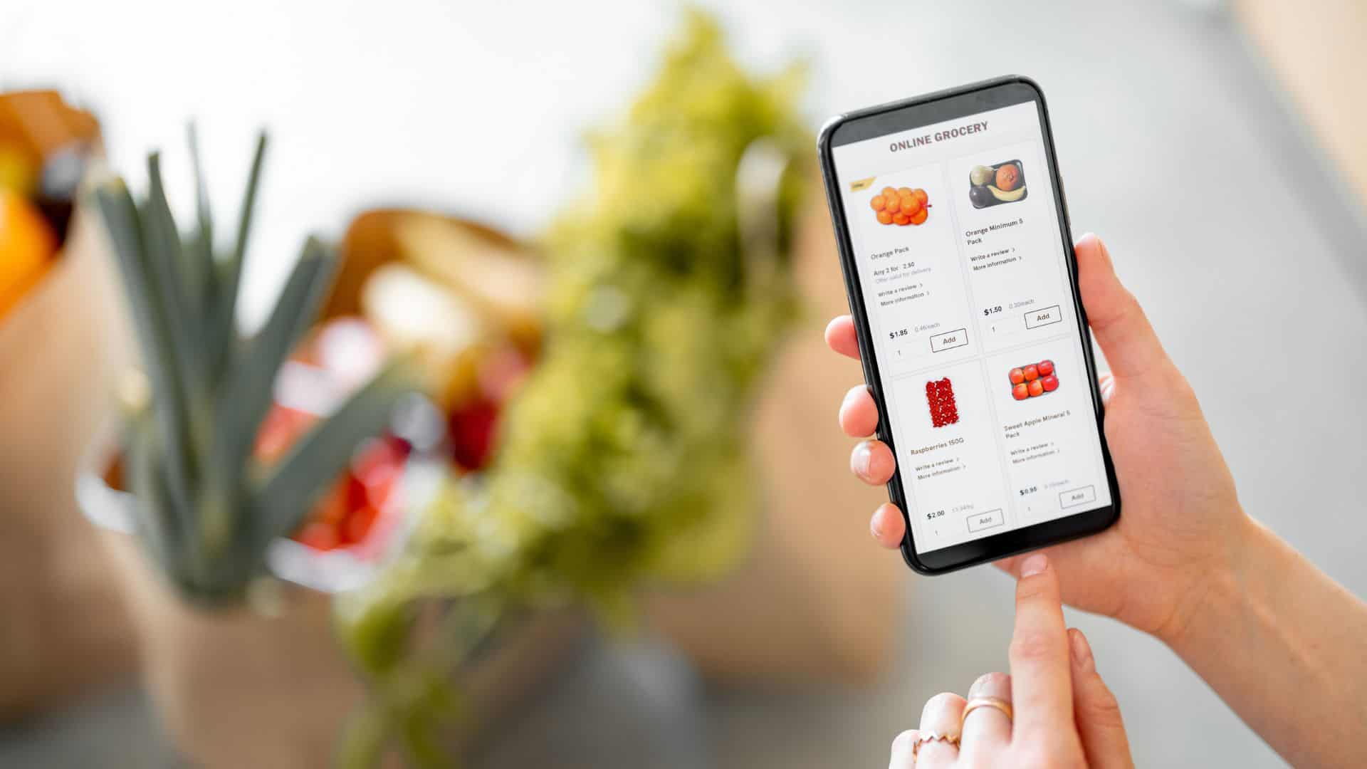 Holding phone with running e-shop application and bags full of fresh food on the background