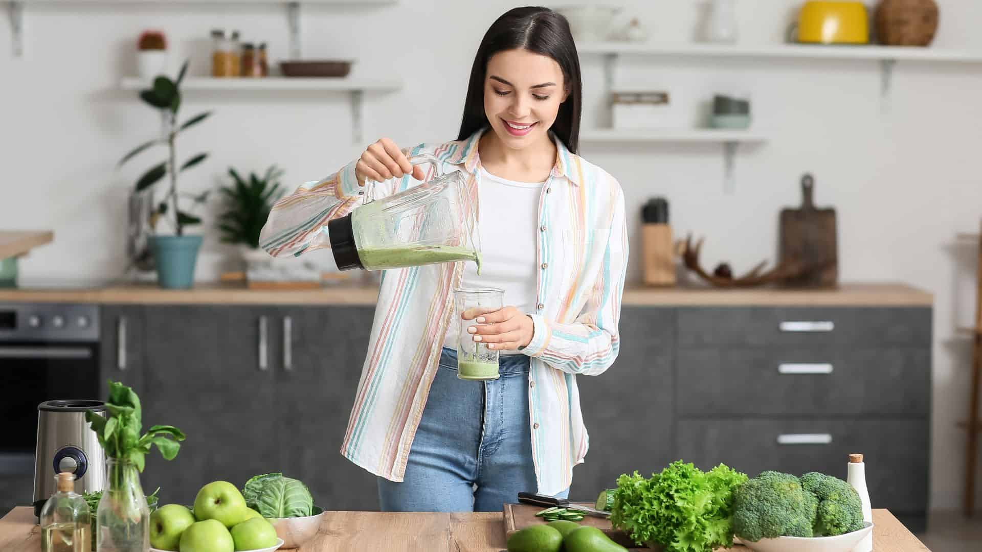 Young woman pouring healthy green smoothie into glass in kitchen